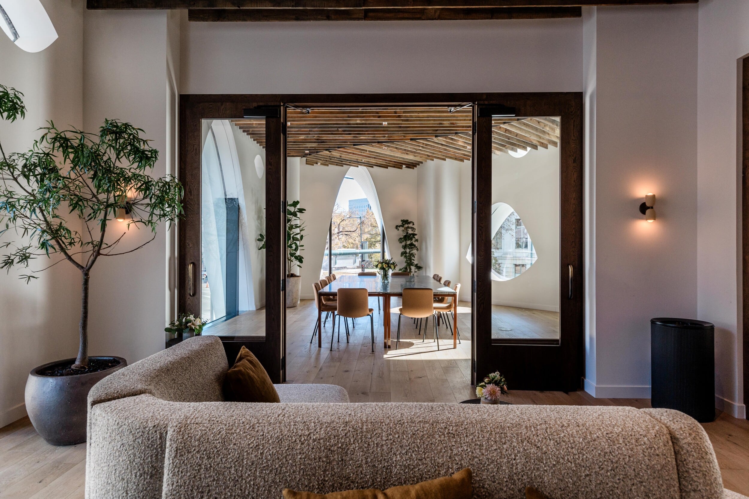 The Overlook meeting space with a cream bouclé sofa and potted tree, looking through open wooden doors into a dining room or meeting space with arched windows and a wood-beamed ceiling.