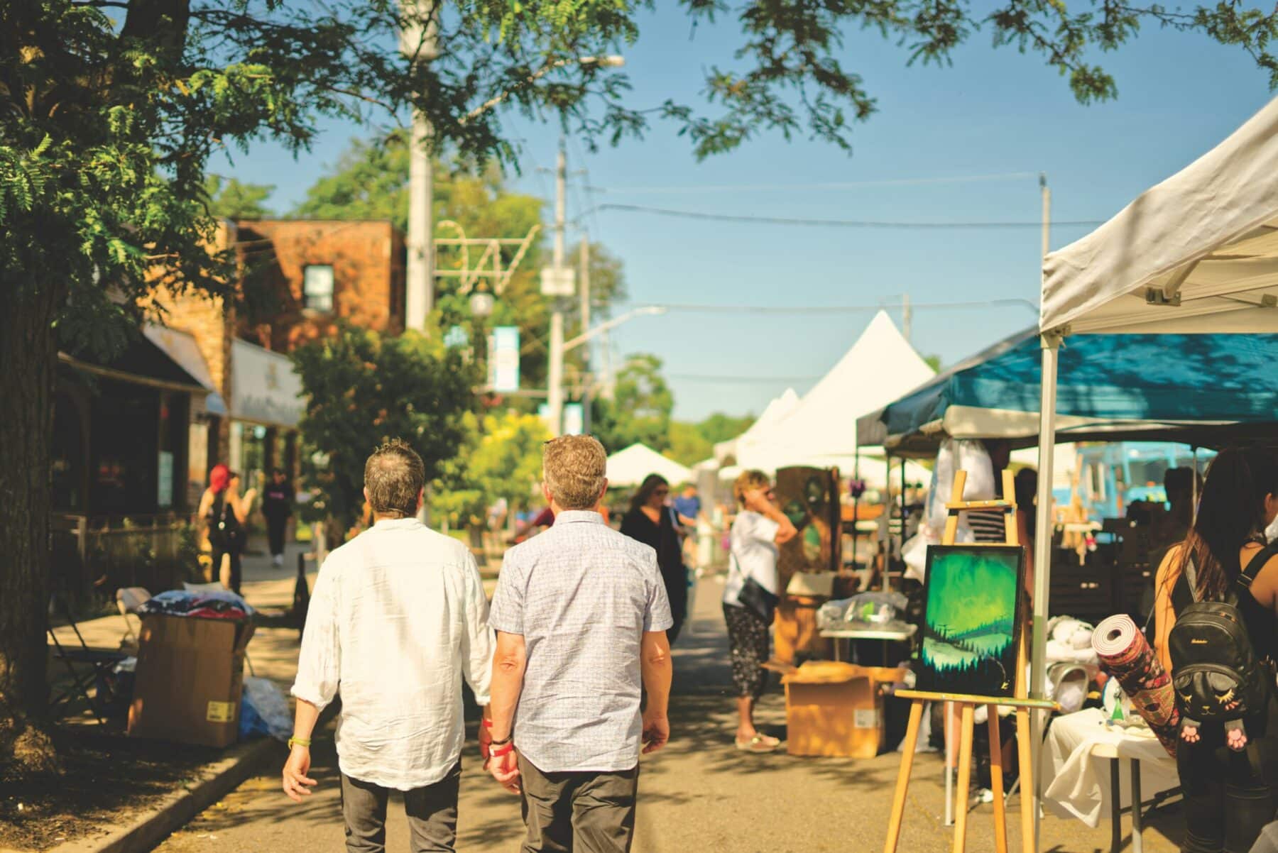 People strolling past white tents and vendor stalls at a sunny outdoor street market.