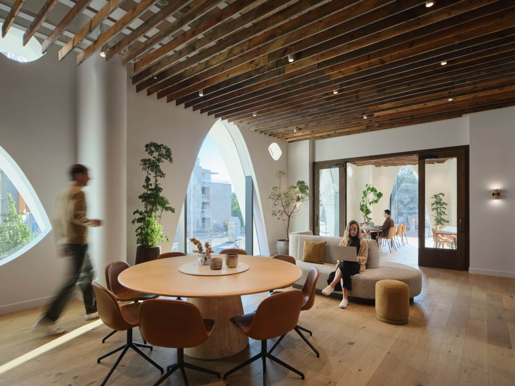 Sunlit level 02 lounge with a round wooden table and leather chairs under a wood-beamed ceiling, with a guest working on a laptop by arched windows and another guest sitting on the couch.