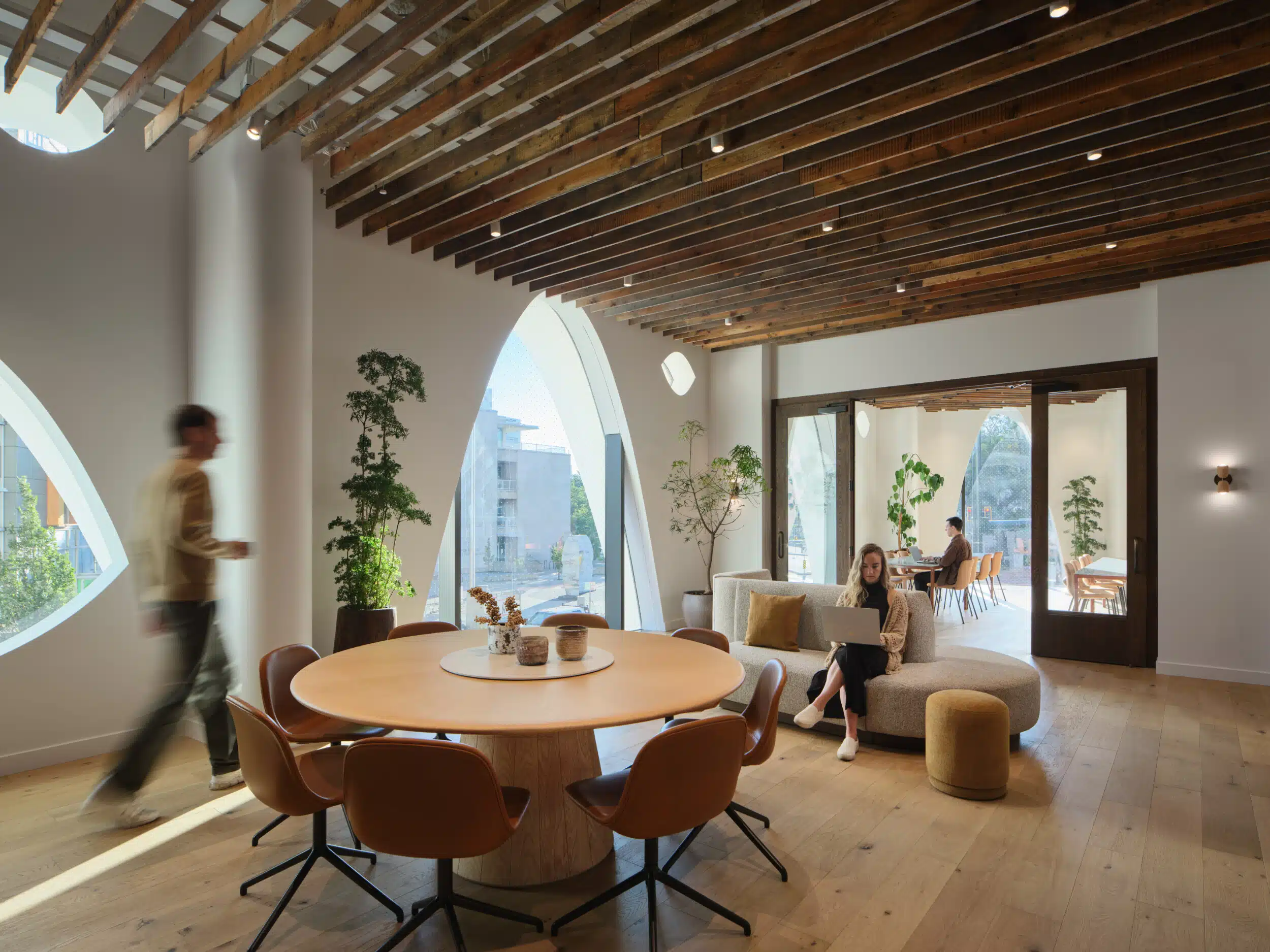 Sunlit level 02 lounge with a round wooden table and leather chairs under a wood-beamed ceiling, with a guest working on a laptop by arched windows and another guest sitting on the couch.