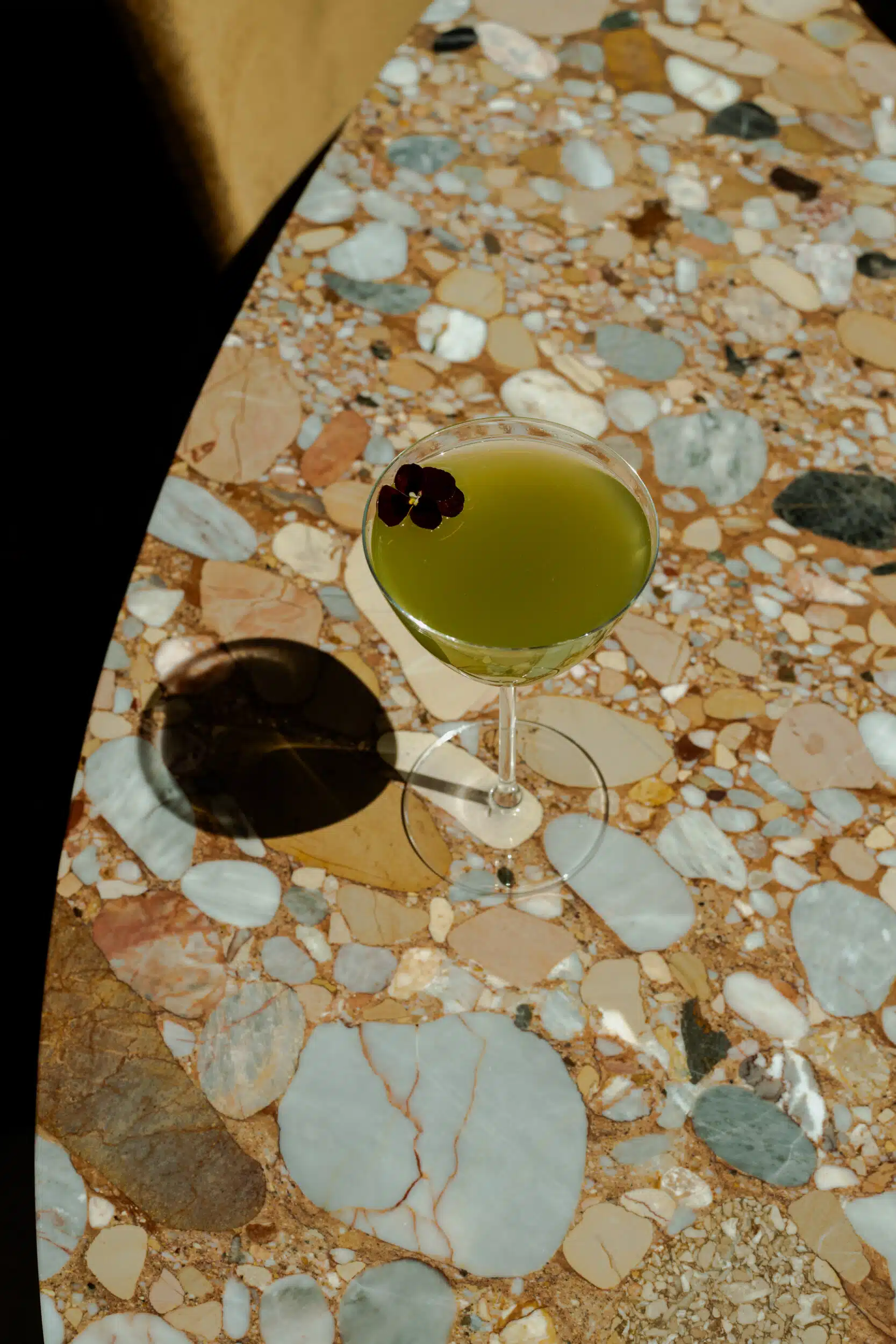 A green cocktail in a coupe glass with a floral garnish, resting on a colorful stone-inlay tabletop at Pasque.