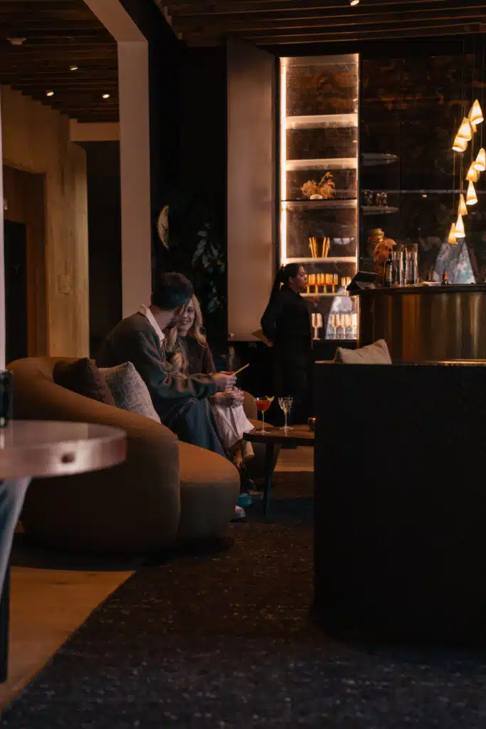 Couple chatting with cocktails on a curved sofa in a dimly lit lounge, with a bartender and server at a brass bar beneath cone-shaped pendant lights.