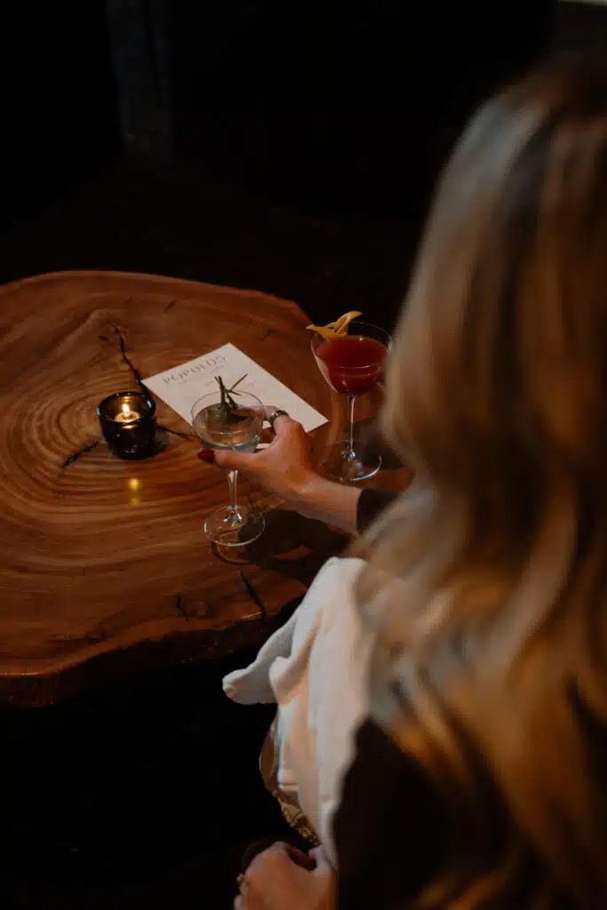 Guest reaching for a rosemary-garnished cocktail at a candlelit wood table, with a red cocktail and the event menu nearby