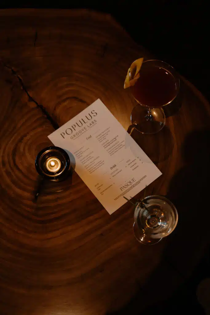 Overhead view of a “Populus Groove Labs” menu beside a candle and two cocktails, one garnished with citrus peel and one with rosemary, on a live-edge wood table.