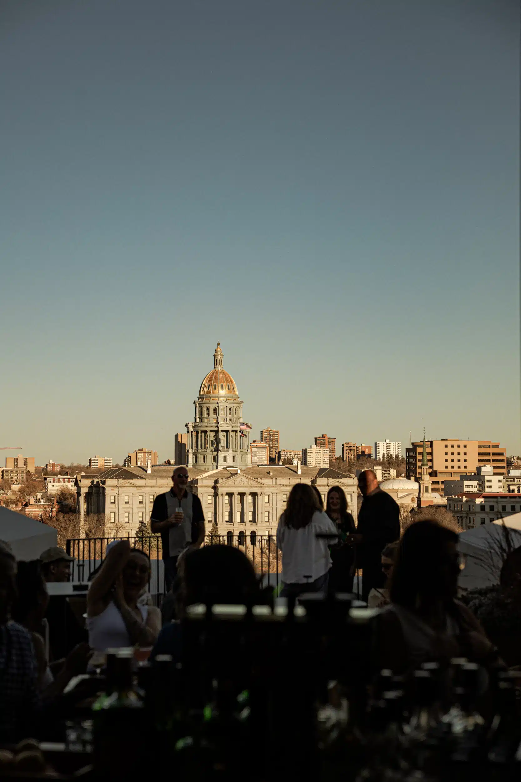 Guests in silhouette mingling with drinks on Stellar Jay's rooftop terrace, with the Colorado State Capitol’s gold dome glowing in late-afternoon sun.