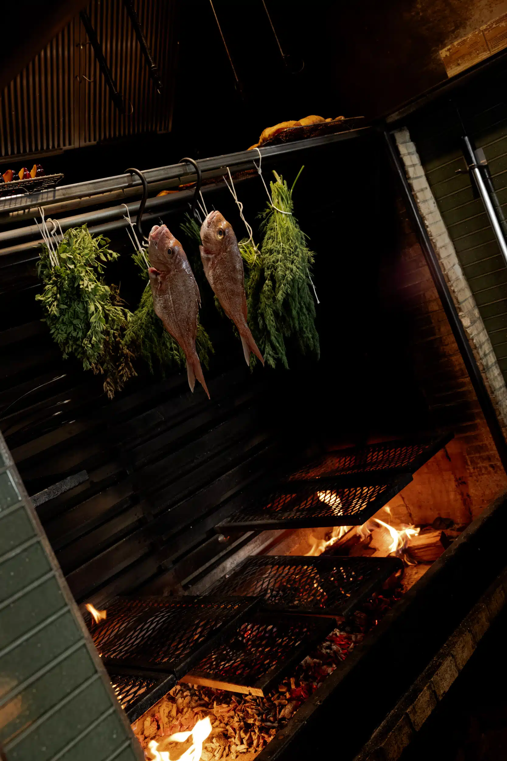 Whole fish and bundles of fresh herbs hanging over a glowing wood-fired hearth at Stellar Jay