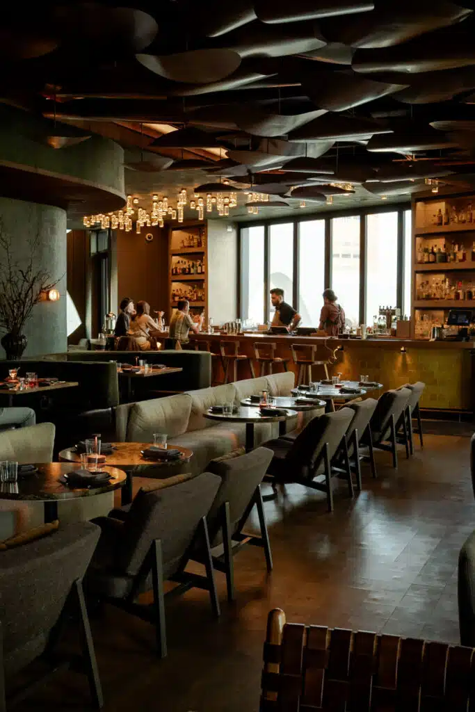 Dimly lit Stellar Jay restaurant with banquette seating, a bar lined with bottles and string lights, and guests seated by tall windows
